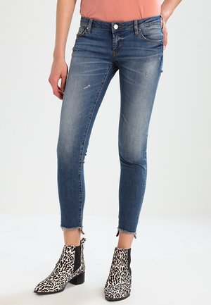 Slim-fit blauwe jeans met lichte slijtage en rafelige zomen, gecombineerd met enkellaarsjes met luipaardprint, gedragen door een persoon met een zalmkleurige top.