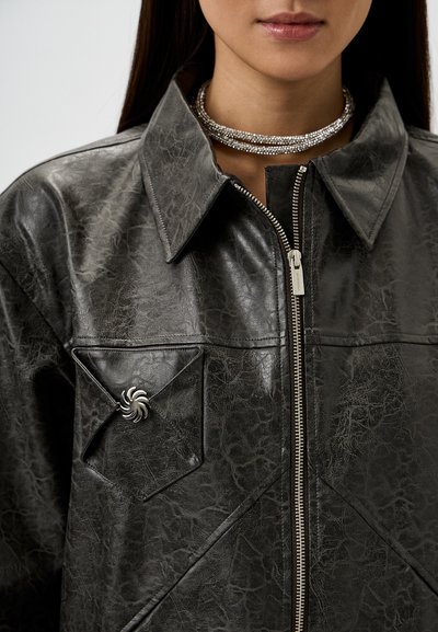 Schwarze Lederimitatjacke mit Reißverschluss, spitzem Kragen, Fronttaschenverzierung und silbernem Spiralakzent. Strukturierte Oberfläche.