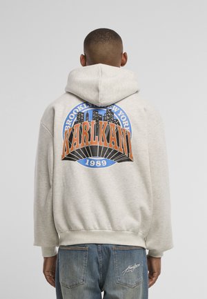 Henkilö, jolla on päällä vaaleanharmaa huppari, jossa on teksti "Brooklyn New York KARLKANI 1989", ja siniset farkut, joissa on allekirjoitus takataskussa, nähtynä takaa.