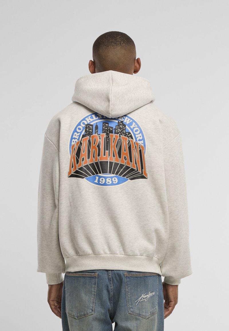 Personne portant un sweat à capuche gris clair avec le motif « Brooklyn New York KARLKANI 1989 » et un jean bleu avec une signature sur la poche arrière, vue de dos.