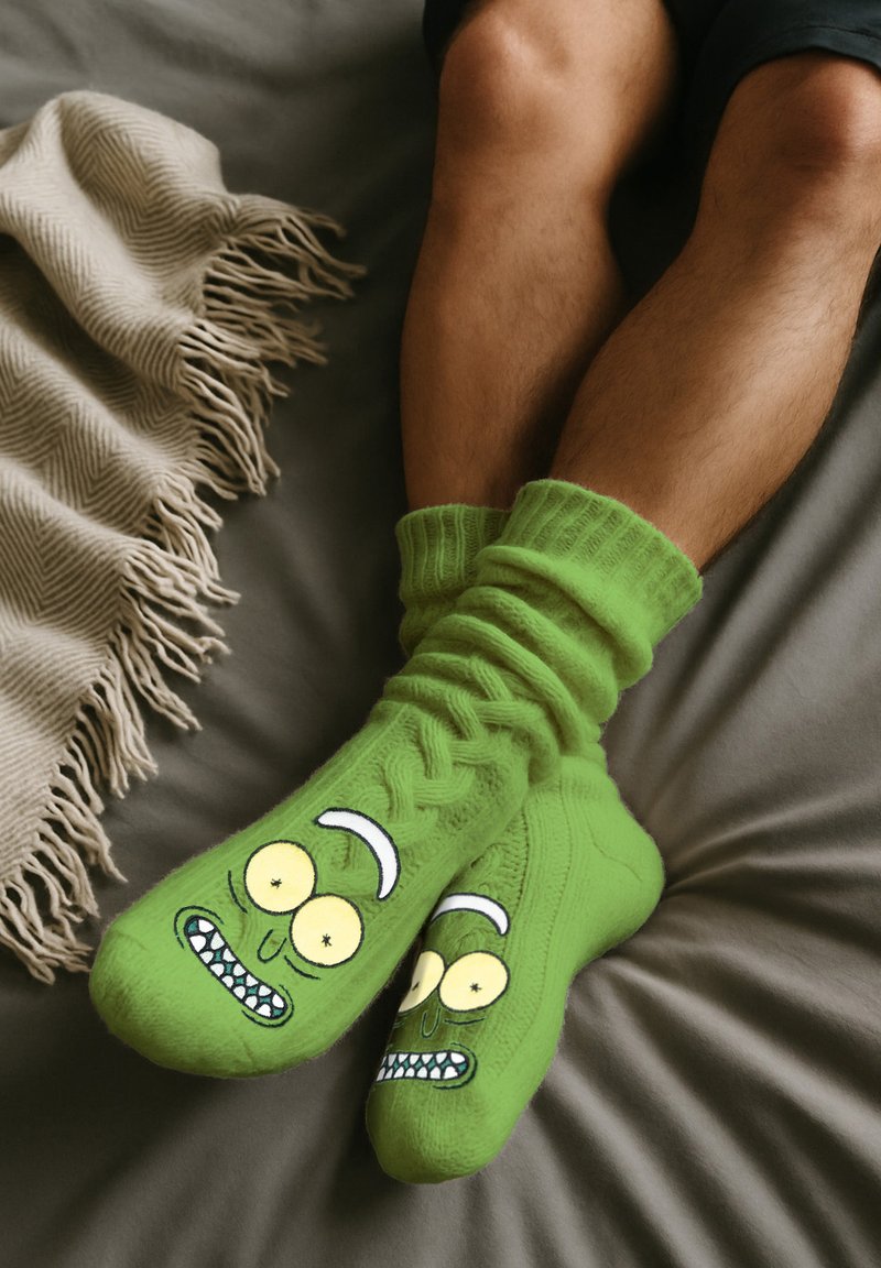 Grüne gestrickte Socken mit einem Cartoon-Gurken-Gesicht, das große Augen und ein zahniges Grinsen zeigt. Getragen an nackten Füßen, mit Rippenstruktur.