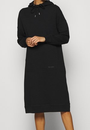 Personne portant une robe noire à capuche à manches longues avec cordon, debout les mains jointes devant, fond clair uni.