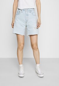 Short en denim bleu clair avec un ourlet effiloché, taille haute et déchirures discrètes. Associé à des baskets montantes blanches.
