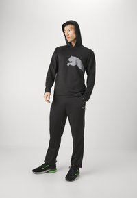 Schwarzer Kapuzenpullover mit großem grauen Puma-Logo, kombiniert mit schwarzen Jogginghosen und schwarzen Sneakers mit grünen Akzenten.