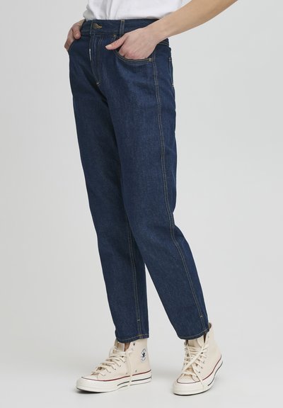 Jeans en denim bleu avec une coupe décontractée, présentant une jambe droite, cinq poches et des coutures jaunes. Portés avec des baskets beiges montantes.