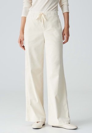 Femme portant un pantalon large en velours côtelé couleur crème avec une taille à cordon et des chaussures blanches sans lacets sur un fond uni.