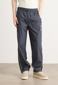 Donker marineblauwe lichtgewicht trainingsbroek met elastische tailleband, zijzakken en witte bies langs de naden. Gestyled met beige sneakers.