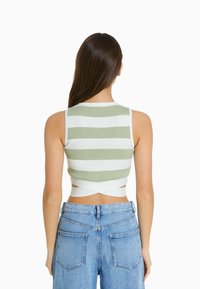 Nainen, jolla on pitkät ruskeat hiukset, pukeutuneena tanktoppiin, jossa on vihreitä ja valkoisia raitoja, ja vaaleansinisiin korkeavyötäröisiin farkkuihin, kääntyy pois.