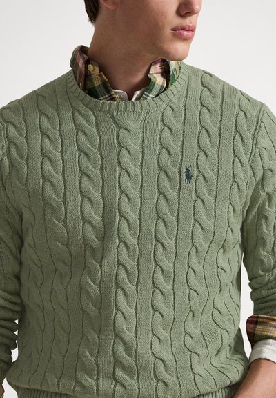 Green cable-knit sweater with round neckline, featuring a small logo on the chest and layered over a plaid shirt with visible collar.