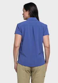Short-sleeve shirt in solid blue, collar style with back pleat detail, made from smooth, lightweight fabric, paired with beige pants.