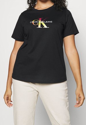 Nainen pukeutunut mustaan Calvin Klein Jeans -t-paitaan, jossa on lyhyet hihat ja värikäs logo, yhdistettynä vaalean beigen sävyisiin housuihin, seisoo yksiväristä taustaa vasten.