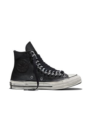 Zapatilla de cuero negra de caña alta con cordones negros gruesos y ojales de metal plateados en cadena, suela de goma blanca con rayas negras y detalles desgastados.