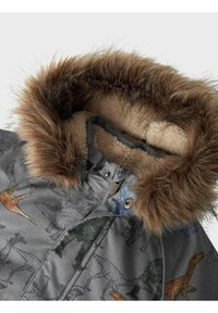 Grå jakke med blødt sherpa-fór, der har en hætte med pelskant. Illustrerede dinosaurer i forskellige farver pryder stoffet.