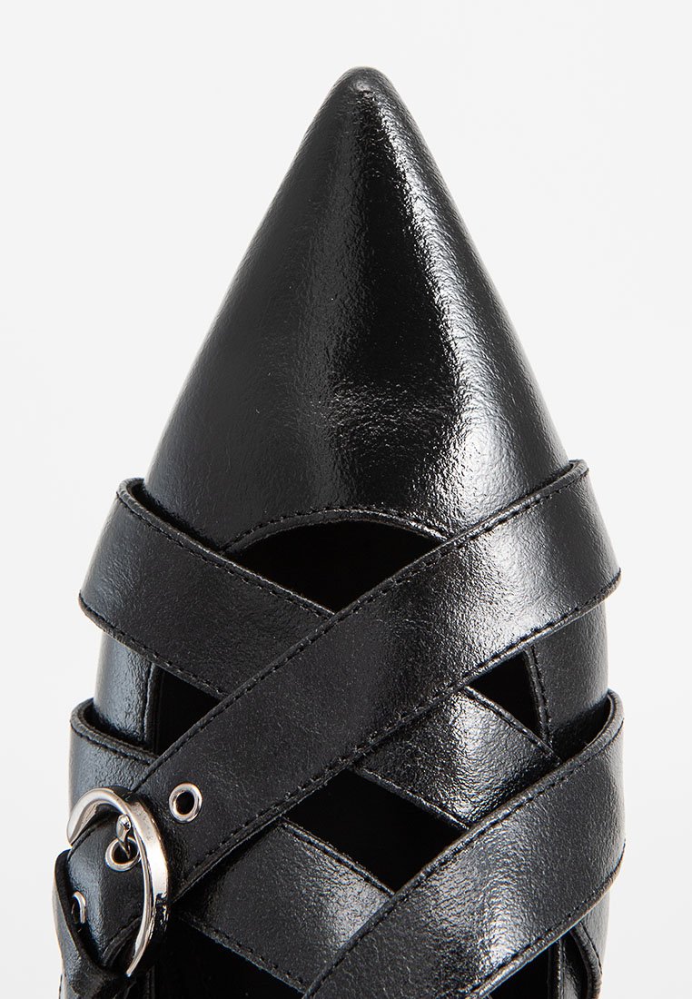 Zapato de cuero negro con punta afilada, con tiras entrecruzadas y un detalle de hebilla plateada sobre un fondo blanco.