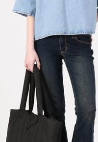 Μαύρη τσάντα tote με λεία υφή, χοντρές λαβές και μοντέρνες ραφές. Φοριέται με ανοιχτόχρωμο τζιν μπλουζάκι και σκούρο τζιν.