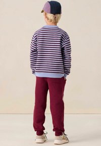 Un pull rayé en violet et bleu clair, associé à un pantalon de survêtement bordeaux. Des accents blancs sur le pantalon et des baskets claires complètent la tenue.