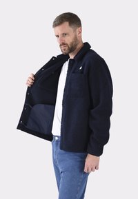 Granatowa polarowa kurtka z teksturowanym wzorem, zapięciem na napy i kieszenią na piersi. Nosi się ją rozpiętą na białej koszuli i niebieskich dżinsach.
