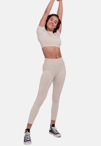 Beige cropped top and high-waisted leggings made of stretchy fabric; model wearing black high-top sneakers, arms raised in an athletic pose.