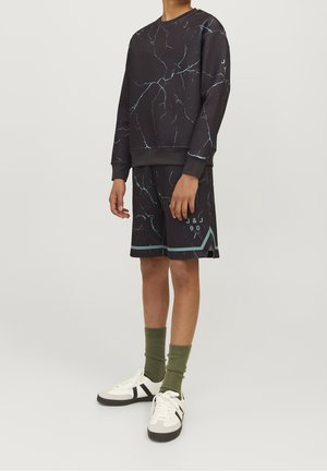 Person iført sort hættetrøje og matchende shorts med blå revnemønster, grønne sokker og hvide sneakers med sorte striber.
