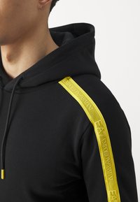 Felpa nera con cappuccio, caratterizzata da un nastro giallo testurizzato sulla spalla con branding visibile, coulisse regolabile e un aspetto morbido del tessuto.