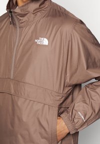Brązowa bluza z kapturem, wykonana z lekkiego materiału, z zamkiem przy szyi, kieszenią na piersi, logo "The North Face", elastycznymi mankietami oraz brandingiem Windwall na rękawie.