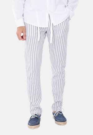 Uomo che indossa pantaloni a righe verticali grigie e bianche, scarpe blu con i lacci e camicia bianca a maniche lunghe, mani nelle tasche.