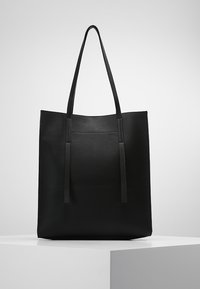 Svart läder tote-väska med långa handtag, rektangulär form och en smal ytterficka, utställd på en vit sockel mot en grå bakgrund.