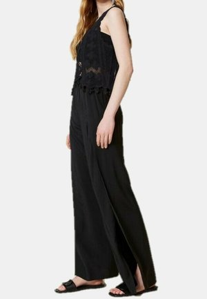 Nainen, joka pukeutunut mustaan hihattomaan virkattuun toppiin, leveälahkeisiin mustiin housuihin, joissa on sivuhalkiot, ja mustiin liukusandaaleihin, seisomassa sivuttain valkoista taustaa vasten.