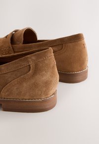 Paire de mocassins en daim marron avec des détails cousus et des petits talons en bois sur un fond clair.