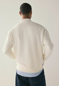 Cream-colored sweatshirt with a ribbed collar and cuffs, featuring a smooth texture and minimalist design, worn over a light blue shirt.