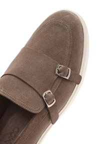Brown suede slip-on shoe with a double buckle strap, white sole, and stitching detail along the edges. Smooth texture and rounded toe.