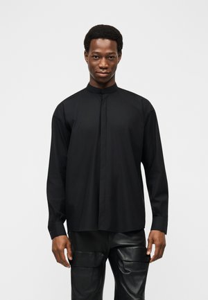 Mężczyzna ubrany w czarną koszulę z długim rękawem i kołnierzem typu mandarynka oraz czarne skórzane spodnie, stojący na jednolitym białym tle.