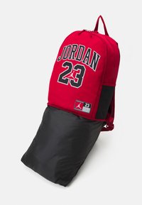 Sac à dos rouge et noir avec "JORDAN 23" en grandes lettres blanches, fermeture zippée, texture lisse et étiquette de logo.