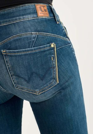 Jean en denim bleu foncé avec une patch en cuir, des accessoires dorés, et un design de poche arrièreDistinctif avec un motif en zigzag.