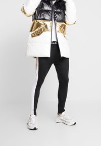 Pěnová bunda s černými, bílými a metalickými zlatými částmi; zapínání na zip, boční kapsy. Černobílé joggers kalhoty s úzkým střihem; šedé tenisky.