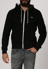 Zwarte zip-up hoodie met een trekkoordcapuchon, contrasterende witte rits en klein logo op de borst. Gemaakt van zachte stof.