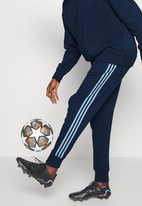 Tuta navy con strisce azzurre chiaro lungo i lati, abbinata a scarpini da calcio neri. Una palla da calcio è in movimento vicino al piede.