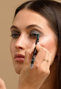 Crayon eyeliner noir et teal appliqué sur l'œil, avec un fard à paupières bleu-gris sur la paupière. Le modèle a de longs cheveux foncés et raides.