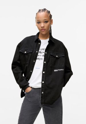Black denim shirt with snap buttons, dual chest pockets, and embroidered logo. Worn over a white graphic t-shirt and gray jeans.