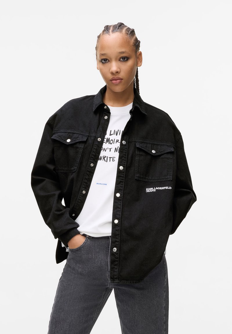 Black denim shirt with snap buttons, dual chest pockets, and embroidered logo. Worn over a white graphic t-shirt and gray jeans.