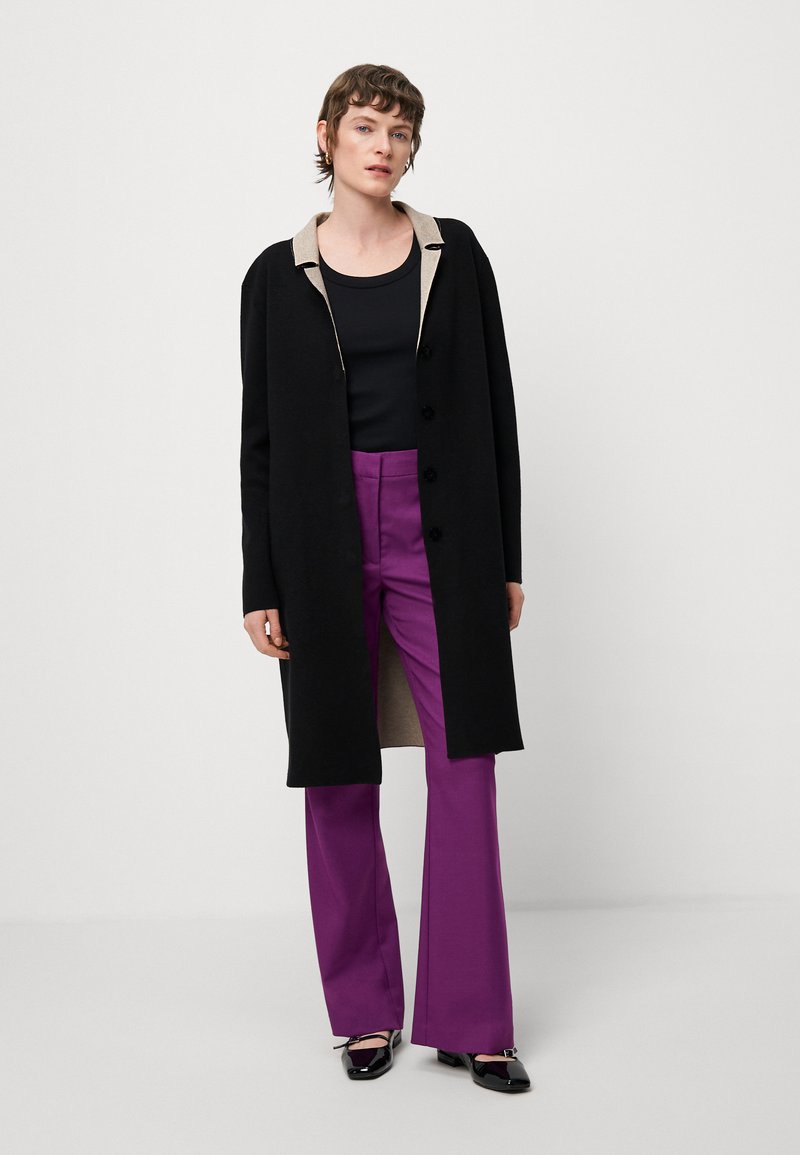 Lungo cappotto nero con colletto beige, bottoni neri, canotta nera e pantaloni viola a zampa d'elefante, abbinati a scarpe nere lucide.