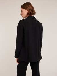 Zwarte blazer met een gestructureerd ontwerp, gelapte revers en manchetten met één knoop. Gladde textuur en op maat gemaakte pasvorm. Achtersteemdetail zichtbaar.