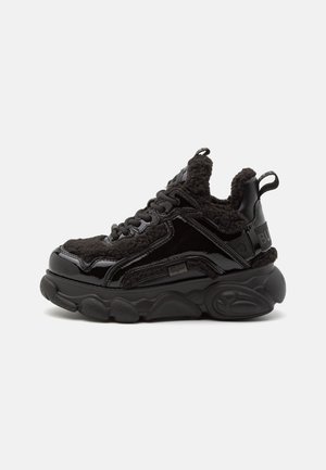Zapatilla negra gruesa con suela gruesa y texturizada, cierre de cordones frontal y forro suave de forro polar visible alrededor del cuello y la lengüeta.