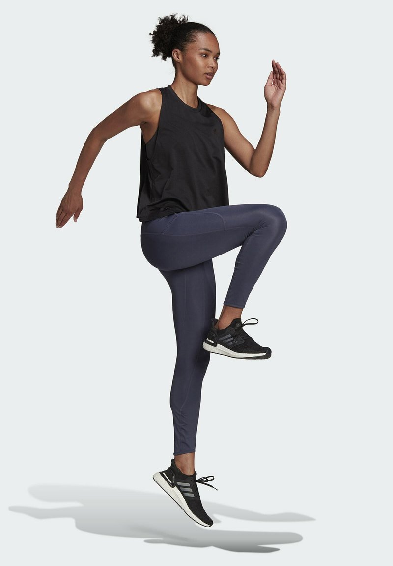 Zwarte mouwloze sporttop, marineblauwe leggings en zwarte hardloopschoenen met witte zolen. Model toont dynamische beweging tegen een lichte achtergrond.