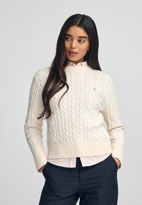 Gebreide crèmekleurige trui met kabelpatronen en geribbelde zoom, gedragen over een lichtroze geblokt shirt. Model poseert tegen een neutrale achtergrond.