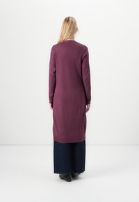 Vrouw met lang blond haar, gekleed in een lange paarse cardigan over donkere wijde broek en zwarte schoenen, staand met haar gezicht naar een witte muur.