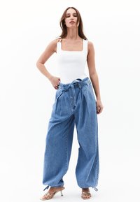 Bodysuit blanco con escote cuadrado y tirantes anchos, combinado con pantalones de mezclilla azul de tiro alto y holgados, que presentan un detalle de cinturón atado. Sandalias doradas de tacón.