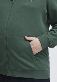 Felpa con zip verde caratterizzata da una texture liscia, polsini a costine e tasche laterali. Accentuata da un logo sul petto e una cerniera sicura.