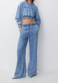 Sudadera recortada de mezclilla azul claro con un bolsillo frontal y cinturilla elástica, combinada con pantalones de pierna ancha a juego que cuentan con un cordón ajustable.