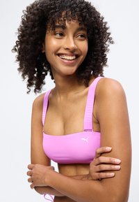 Γυναίκα με σγουρά μαλλιά που χαμογελά, φοράει ροζ αθλητικό σουτιέν Puma, με σταυρωμένα τα χέρια, κοιτάζει προς τα πλάγια μπροστά σε λευκό φόντο.